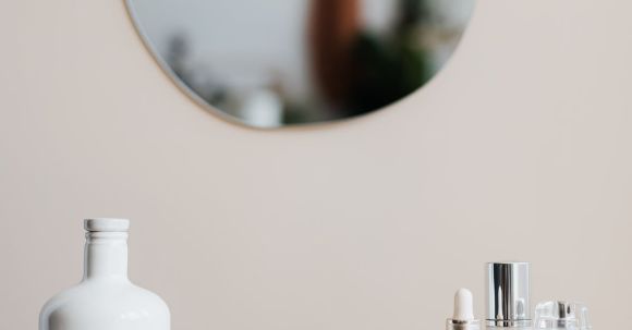 Storage Hacks - Reusable cosmetic containers for cream and shower gel arranged near various skincare products and perfume bottle on white marble shelf against beige wall with mirror in elegant bathroom interior